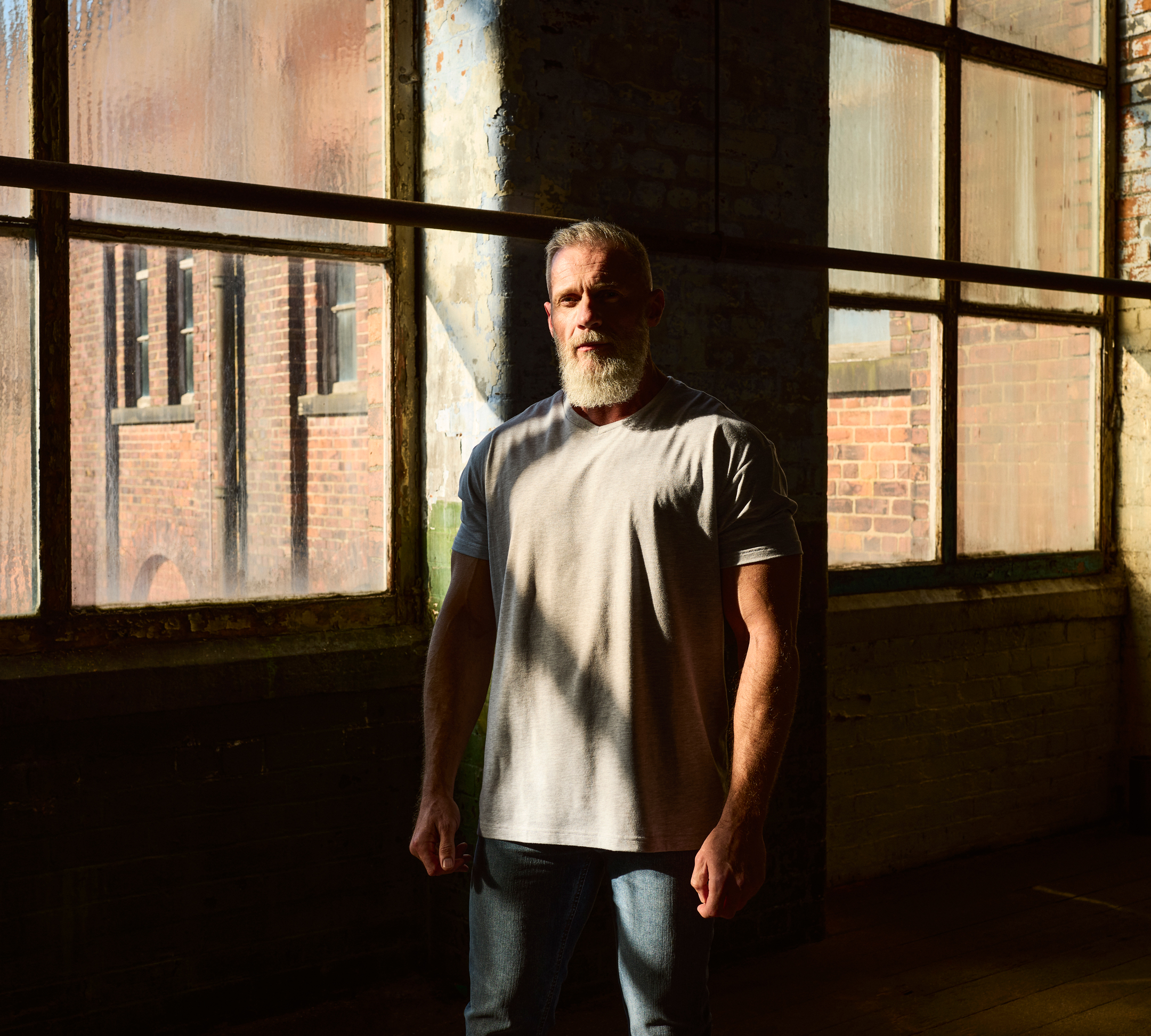 Bearded male model wearing a casual grey t-shirt and jeans, photographed in natural light against an industrial brick backdrop for a clothing brand campaign